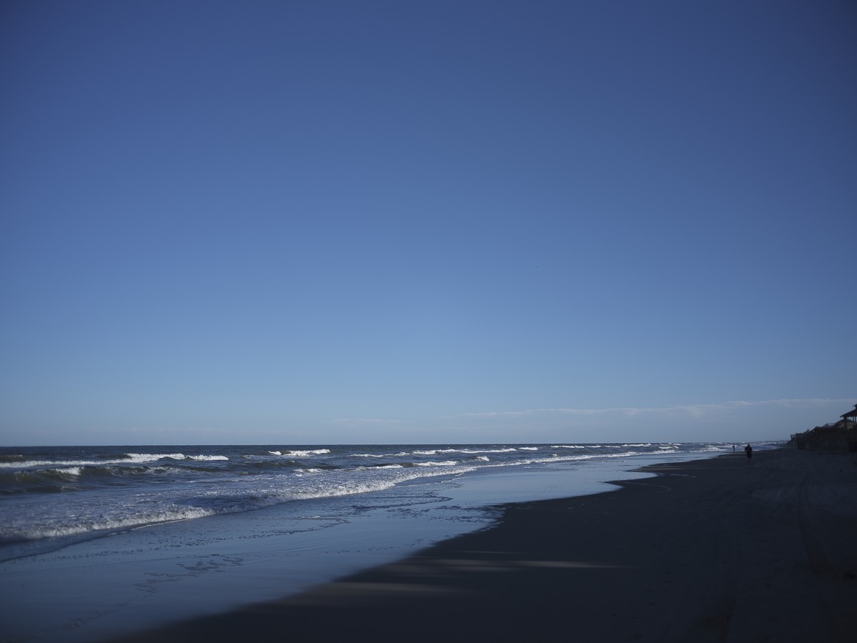 An empty beach with breaking waves under a huge deep-blue sky, one distant figure at the water's edge.