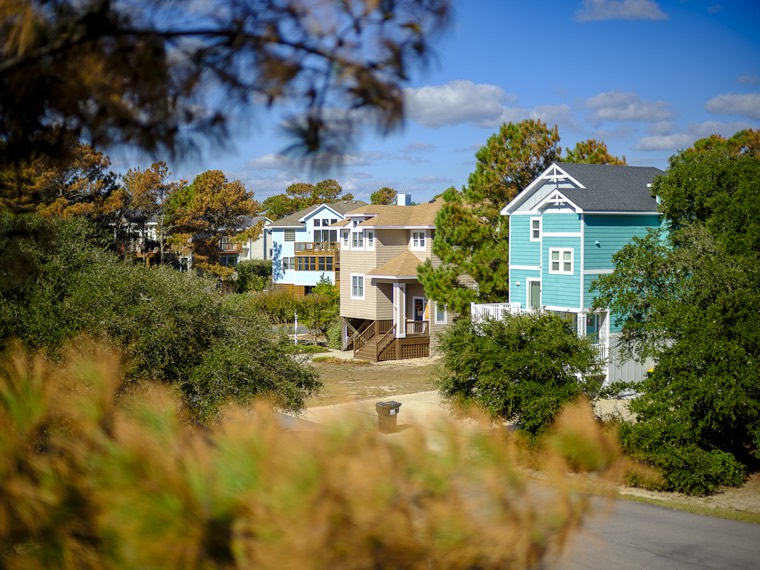 Pastel beach houses, one aqua and one tan, framed by pine branches under a blue sky.