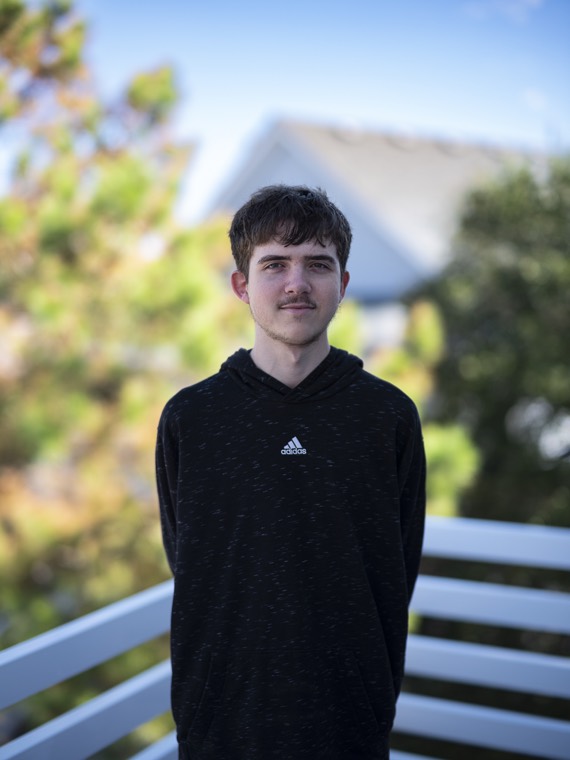 A teenager in a black hoodie standing at a white deck railing with a house roof and pines behind.