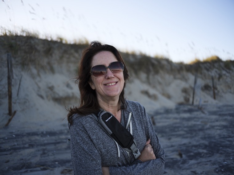 A smiling person in sunglasses and a grey hoodie, arms crossed, in front of grassy dunes.