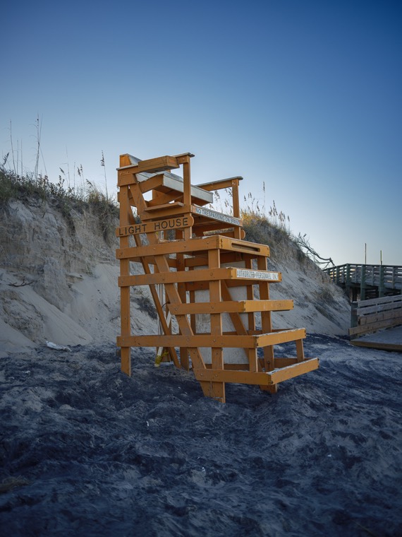 A wooden lifeguard stand labelled Light House on an empty beach at dusk, dunes and sea oats behind it.